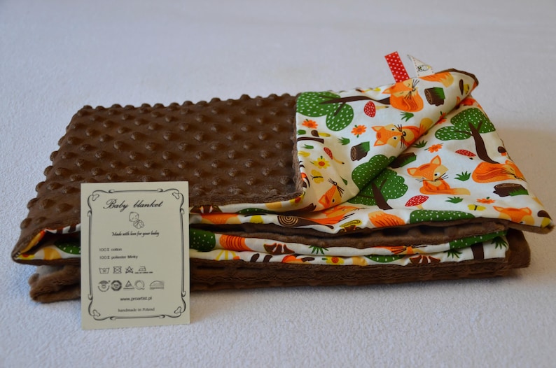 May include: A brown and white baby blanket with a fox print. The blanket is made of soft, plush fabric and has a textured surface. The blanket is folded in half and has a small tag attached to it that says "Baby blanket".