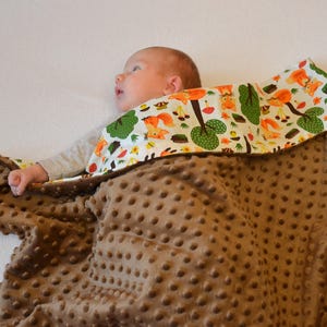 May include: A brown baby blanket with a white and green patterned fabric on top. The blanket is made of a soft, plush material with a textured surface. The patterned fabric features a design of trees, squirrels, and other woodland creatures.