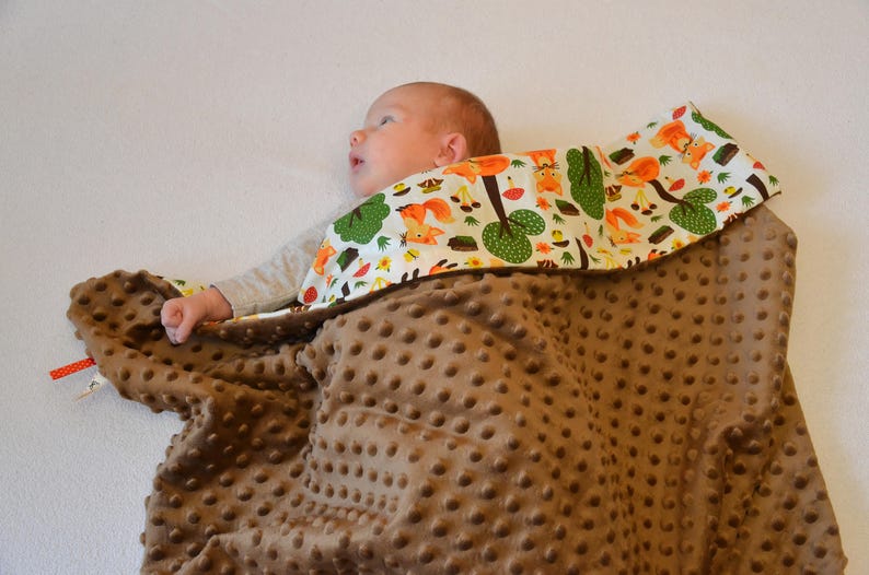 May include: A brown baby blanket with a white and green patterned fabric on top. The blanket is made of a soft, plush material with a textured surface. The patterned fabric features a design of trees, squirrels, and other woodland creatures.