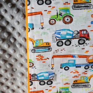 May include: A white fabric with a repeating pattern of construction vehicles. The vehicles are drawn in a cartoon style and include a cement mixer, a crane, a bulldozer, and a tractor. The background is white with a few orange bricks.