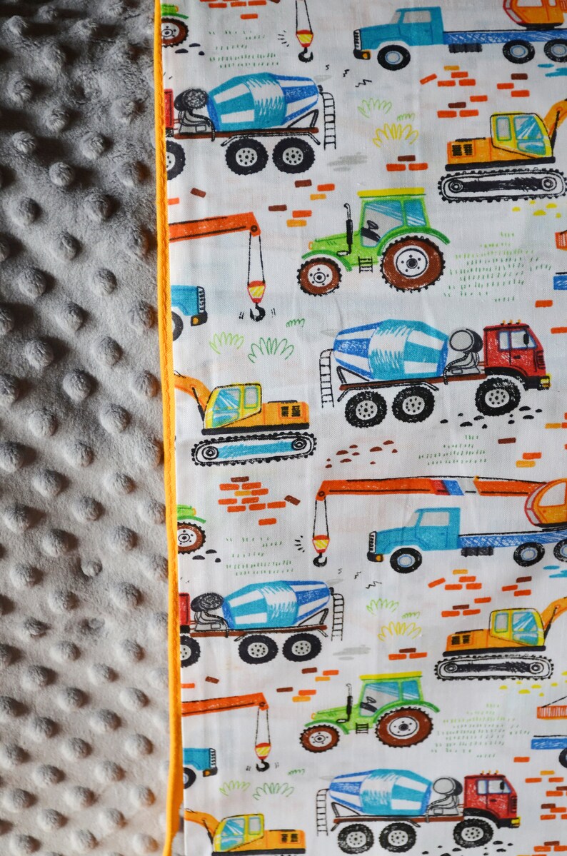 May include: A white fabric with a repeating pattern of construction vehicles. The vehicles are drawn in a cartoon style and include a cement mixer, a crane, a bulldozer, and a tractor. The background is white with a few orange bricks.