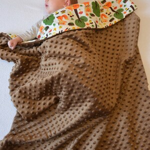 May include: A brown baby blanket with a textured surface. The blanket is made of soft, plush fabric and has a white and green patterned lining with a woodland animal design.