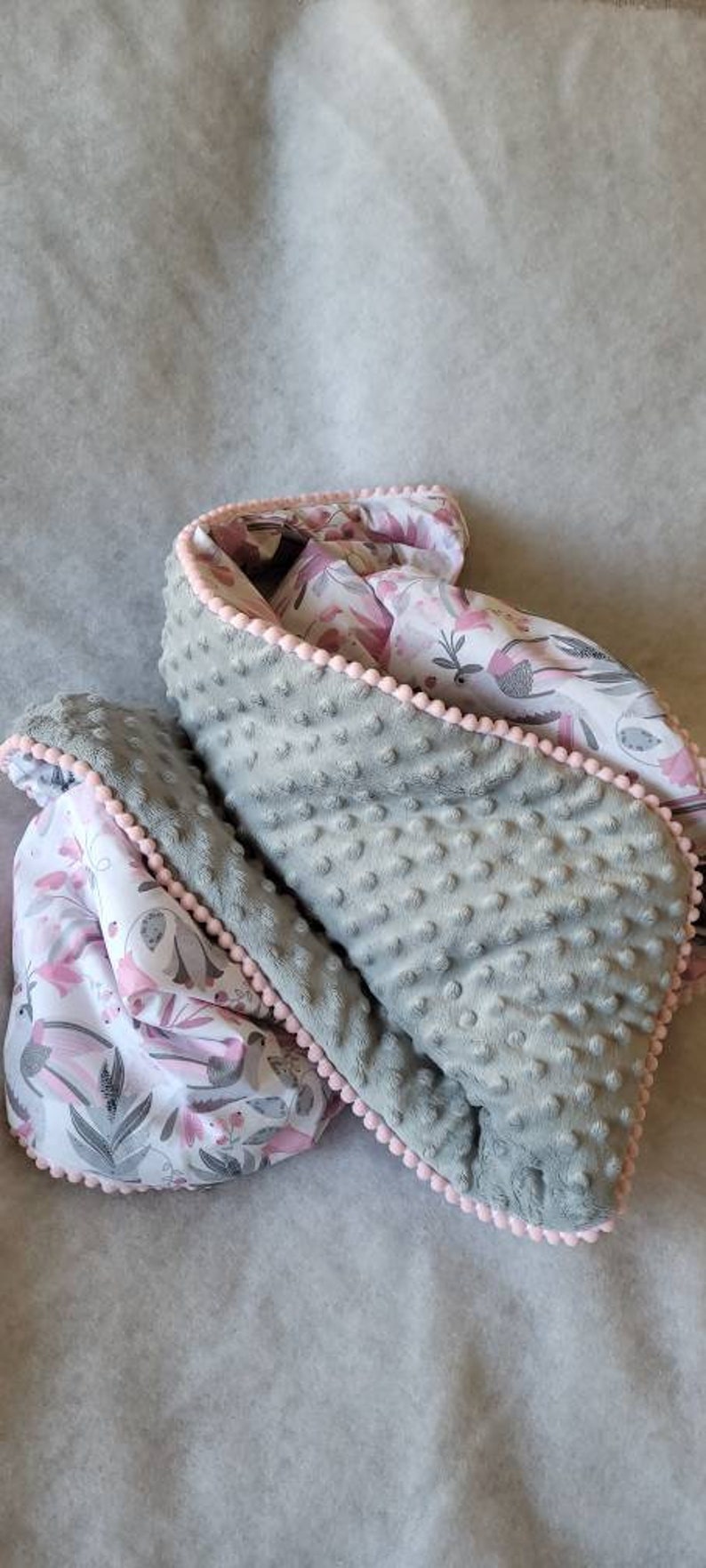 Pode incluir: Um cobertor de beb&ecirc; cinza e branco com uma borda de pompom rosa. O cobertor &eacute; feito de tecido macio e felpudo e apresenta um padr&atilde;o floral.