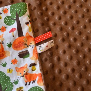 May include: A white fabric baby blanket with a fox and forest print on one side and a brown minky fabric on the other side. The blanket has two fabric tags with an owl design.