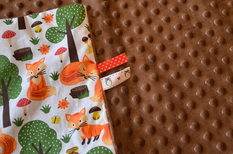 May include: A white fabric baby blanket with a fox and forest print on one side and a brown minky fabric on the other side. The blanket has two fabric tags with an owl design.