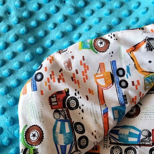 May include: A turquoise blue minky dot blanket with a white cotton fabric backing. The cotton fabric features a repeating pattern of construction vehicles, including cement mixers, dump trucks, and excavators. The fabric is trimmed with a yellow border.