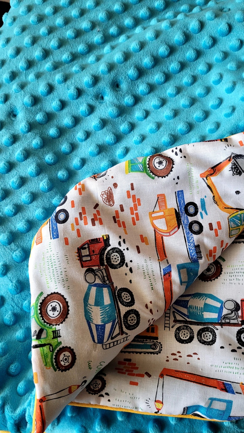 May include: A turquoise blue minky dot blanket with a white cotton fabric backing. The cotton fabric features a repeating pattern of construction vehicles, including cement mixers, dump trucks, and excavators. The fabric is trimmed with a yellow border.