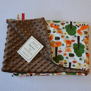 May include: A brown and white baby blanket with a fox and forest design. The blanket has a soft, textured side and a printed side. The tag reads "Baby Blanket Handmade with Love".