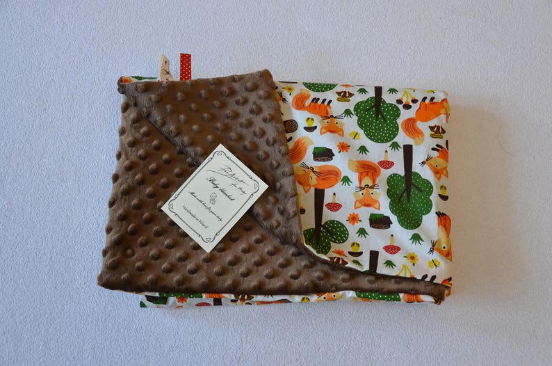 May include: A brown and white baby blanket with a fox and forest design. The blanket has a soft, textured side and a printed side. The tag reads "Baby Blanket Handmade with Love".