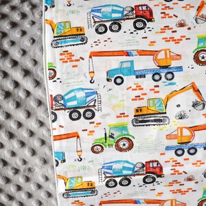 May include: White fabric with a repeating pattern of construction vehicles, including cement mixers, excavators, and cranes. The vehicles are illustrated in bright colors, including yellow, orange, blue, green, and red. The background is white with a subtle pattern of orange bricks.