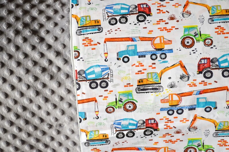 May include: White fabric with a repeating pattern of construction vehicles, including cement mixers, excavators, and cranes. The vehicles are illustrated in bright colors, including yellow, orange, blue, green, and red. The background is white with a subtle pattern of orange bricks.