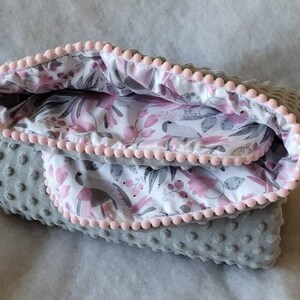 Pode incluir: Um cobertor de beb&ecirc; cinza e rosa com estampa floral e acabamento de pompons rosa.