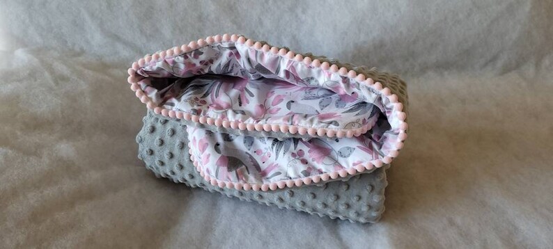Pode incluir: Um cobertor de beb&ecirc; cinza e rosa com estampa floral e acabamento de pompons rosa.