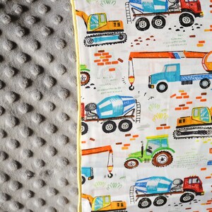 May include: A white fabric with a yellow border features a repeating pattern of construction vehicles. The vehicles are drawn in a cartoon style and include cement mixers, excavators, and cranes. The background is white with a light green and brown pattern.