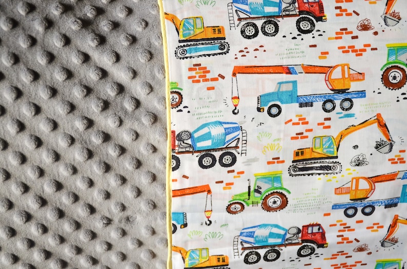 May include: A white fabric with a yellow border features a repeating pattern of construction vehicles. The vehicles are drawn in a cartoon style and include cement mixers, excavators, and cranes. The background is white with a light green and brown pattern.