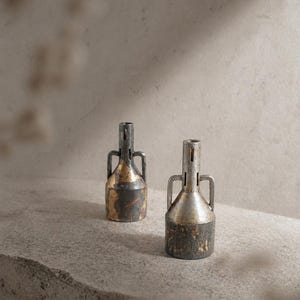Pode incluir: Dois pequenos vasos de metal em estilo vintage com alças. Os vasos têm uma base cilíndrica e um pescoço estreito com uma fenda vertical. O metal tem uma aparência desgastada e envelhecida com tons dourados e marrom escuro.
