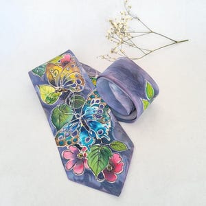 Puede incluir: Una corbata con un diseño pintado a mano sobre un fondo morado. El patrón muestra dos grandes mariposas en tonos amarillos, naranjas y azules, rodeadas de hojas verdes y flores rosas. La tela tiene una textura suave y sedosa.