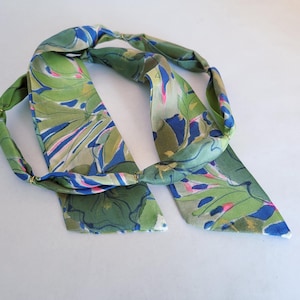May include: A long, narrow fabric hair accessory featuring a vibrant botanical print in shades of olive green, royal blue, and bright pink. The accessory is constructed with small, evenly spaced gold-toned metal rings that segment the fabric.