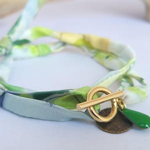 Peut inclure: Un bracelet en soie avec un fermoir à bascule doré et un charm en forme de goutte vert. Le tissu du bracelet présente un motif floral aquarelle dans les tons de vert, jaune et bleu. Un petit charme rond de couleur bronze est également attaché.