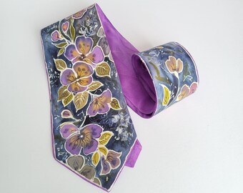 Pode incluir: Uma gravata com um padrão floral pintado à mão com flores roxas, amarelas e brancas sobre um fundo azul escuro. A gravata tem um forro roxo sólido e uma ponta afiada, exibindo uma textura de tecido suave em todo o seu design.