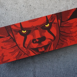 May include: A close-up of a red and black illustration of a clown with yellow eyes. The clown's mouth is open and shows teeth. The background is dark red.