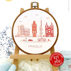 May include: A white embroidery hoop with a red and white illustration of the Prague skyline. The hoop is on a wooden easel and surrounded by craft supplies, including embroidery floss, scissors, and sequins. The text "PRAGUE" is printed below the illustration.