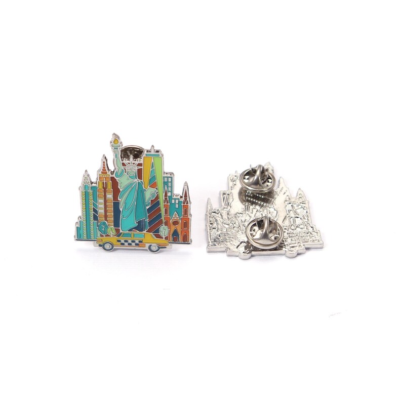 May include: A colorful enamel pin featuring the New York City skyline, including the Statue of Liberty and various skyscrapers. The pin is mounted on a yellow taxi cab. The back of the pin is silver with two pin backs.