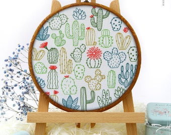 May include: A white fabric hoop with a colourful embroidery design of various cacti and succulents. The hoop is mounted on a wooden easel and surrounded by craft supplies, including blue ribbon, gold sequins, and embroidery floss.