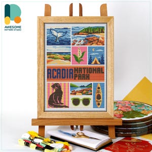 May include: A framed cross-stitch depicting scenes from Acadia National Park. The artwork includes images of a whale tail, coastline, beach, plant, tent, and sunglasses. The text "ACADIA NATIONAL PARK" is prominently displayed. The frame is displayed on a wooden easel.