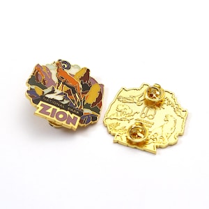 May include: Two gold-colored enamel pins. One pin features a landscape with a ram and the text "ZION NATIONAL PARK". The other pin is a gold-colored outline of a landscape with text.