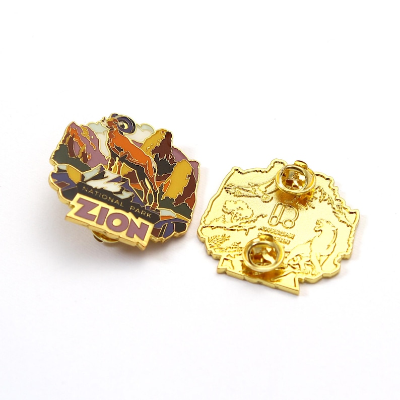 May include: Two gold-colored enamel pins. One pin features a landscape with a ram and the text "ZION NATIONAL PARK". The other pin is a gold-colored outline of a landscape with text.