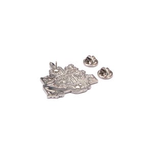 May include: A silver-colored pin shaped like a detailed city map, with two pin backs. The pin features intricate architectural details and is designed to be attached to clothing or accessories. The pin is a collectible item.