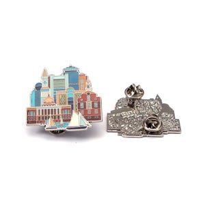 May include: A colourful enamel pin depicting a cityscape with buildings, a clock tower, and sailboats. The pin has a detailed design with various colours and a silver metal backing with a pin clasp.