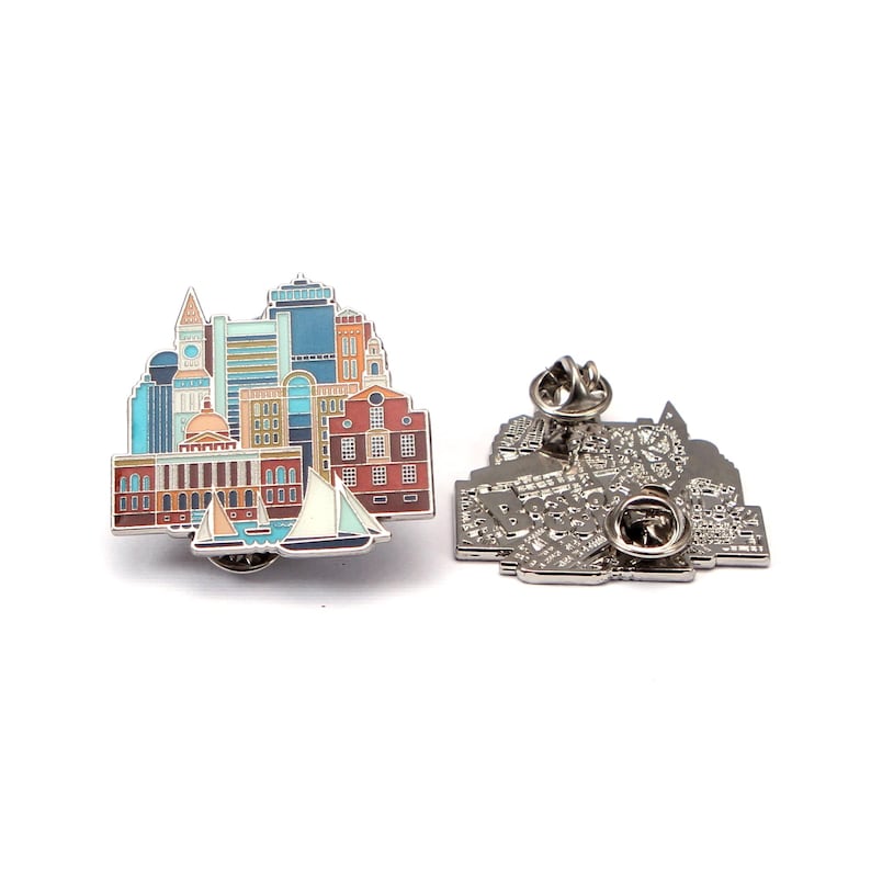 May include: A colourful enamel pin depicting a cityscape with buildings, a clock tower, and sailboats. The pin has a detailed design with various colours and a silver metal backing with a pin clasp.