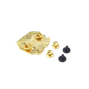May include: A gold-colored metal pin with a detailed design, featuring two pin backs attached and three additional pin backs. The pin is on a white background.