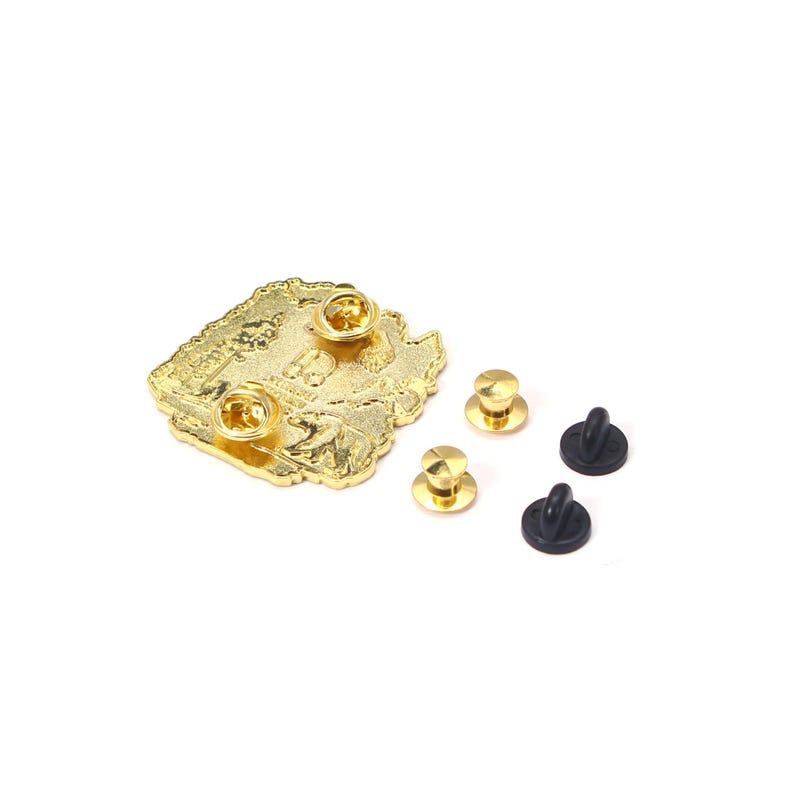 May include: A gold-colored metal pin with a detailed design, featuring two pin backs attached and three additional pin backs. The pin is on a white background.