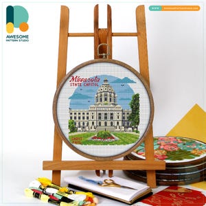 May include: A cross-stitch embroidery of the Minnesota State Capitol building in a wooden hoop, displayed on a wooden easel. The embroidery features the building, green trees, and blue sky. The text "Minnesota State Capitol" and "Saint Paul" are visible.