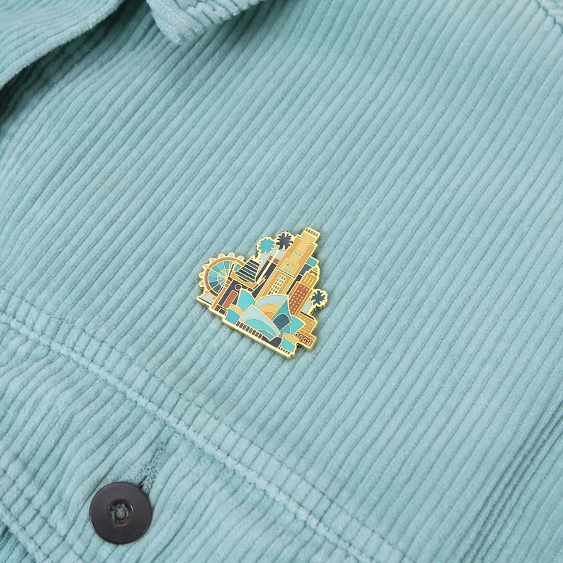 May include: A gold-colored enamel pin featuring a cityscape design with buildings, a Ferris wheel, and palm trees. The pin is attached to a light blue corduroy jacket. The pin has blue, orange, and brown accents.