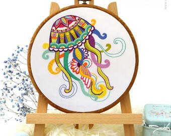 May include: A colorful cross-stitch embroidery of a jellyfish displayed on a wooden easel. The jellyfish design features vibrant hues of blue, green, yellow, and purple. The embroidery hoop is surrounded by craft supplies, including thread, scissors, and a kit.