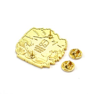 K&ouml;nnte beinhalten: Goldfarbene Emaille-Anstecknadel mit detailliertem Design mit Wellen, einem Vogel und japanischem Text. Die Anstecknadel hat zwei Nadelr&uuml;cken und den Text "AMERICAN PATTERN STUDIO".