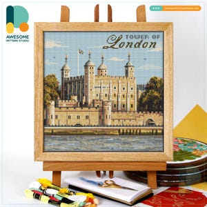 May include: A framed cross-stitch of the Tower of London, displayed on a wooden easel. The artwork features the historic castle in shades of beige and brown, with the text "Tower of London" above. The sky is light blue. Embroidery floss and scissors are in the foreground.