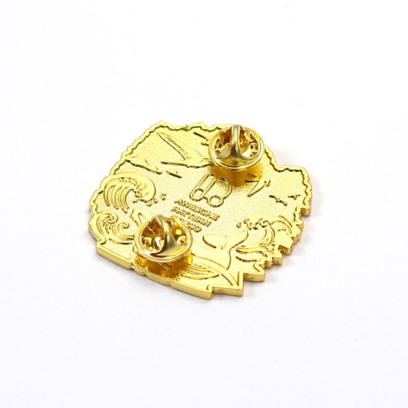 K&ouml;nnte beinhalten: Goldfarbene Anstecknadel mit detailliertem Design mit Wellen, V&ouml;geln und dem Text "AMERICAN GATHERING CO." Die Anstecknadel hat zwei Nadelr&uuml;cken f&uuml;r sicheren Halt. Die Gesamtform ist unregelm&auml;&szlig;ig, mit strukturierter Oberfl&auml;che.