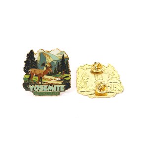 May include: A Yosemite National Park pin featuring a landscape with a mountain, trees, and a ram. The pin has a gold border and the text "YOSEMITE NATIONAL PARK". The back of the pin is gold with two pin backs.