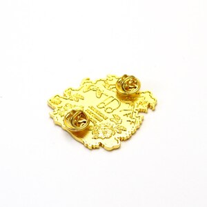 May include: Gold-colored enamel pin with a floral design and the text "AMERICAN PATTERN 1980" on the back. The pin has two butterfly clasps.