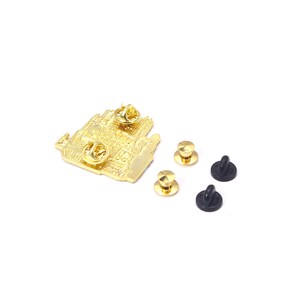 May include: A gold-colored metal pin with a detailed architectural design, featuring two pin backs. Includes three gold-colored pin clutches and two black rubber pin backs. The pin is on a white background.