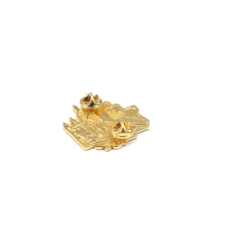 Puede incluir: Un pin de metal dorado con dos cierres de alfiler. El pin tiene un dise&ntilde;o detallado, posiblemente de un edificio o un mapa. El pin est&aacute; sobre un fondo blanco.