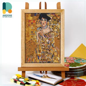 May include: A framed cross-stitch of a woman in a gold and patterned dress, displayed on a wooden easel. The artwork is in a wooden frame. The image includes embroidery floss, scissors, and a notebook. The logo "AWESOME PATTERN STUDIO" is in the upper left corner.