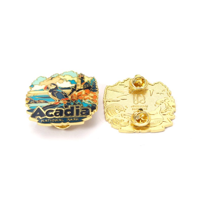 K&ouml;nnte beinhalten: Eine goldfarbene Anstecknadel mit einem detaillierten Emaille-Design mit einem Papageientaucher, einem Leuchtturm und dem Text "Acadia National Park". Die Anstecknadel hat eine abgerundete Form mit goldfarbener R&uuml;ckseite und zwei Nadelverschl&uuml;ssen.