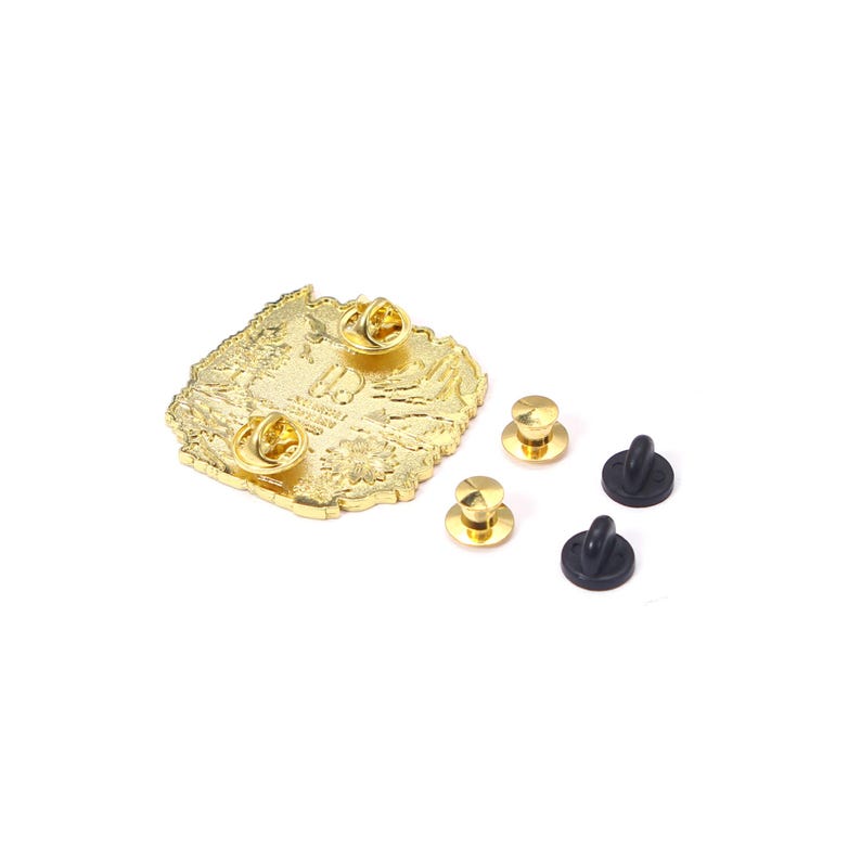 May include: A gold-colored metal pin with a detailed design, featuring three pin backs attached and three additional pin backs and four black rubber clutches. The pin has a textured surface and a unique shape.