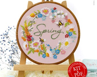 May include: A pink embroidery hoop featuring a floral wreath and the word "Spring" in cursive. Two bees are embroidered within the wreath. The hoop is displayed on a wooden stand. The image also includes a blue ribbon, a small box, and a set of embroidery floss.